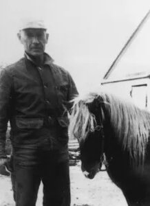 Black and white photograph of an older man in a work jacket and cap standing next to a small pony with a long mane, set against a rustic background.