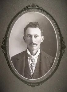 Black and white portrait of a man from the early 1900s, featuring a mustache and wearing a formal suit with a tie, framed in an ornate oval frame.
