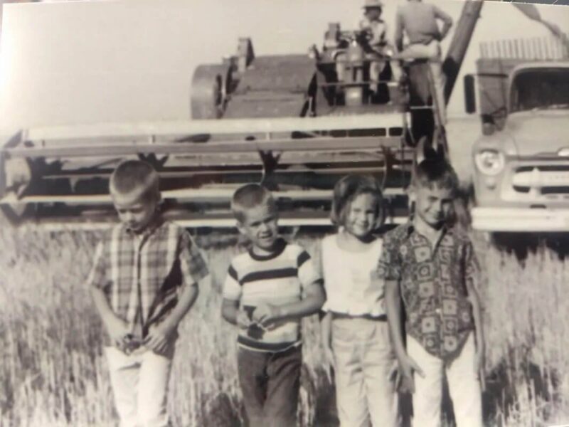 Black and white photograph of four children standing in a field with a vintage farm truck and harvesting equipment in the background, showcasing rural life in the mid-20th century.