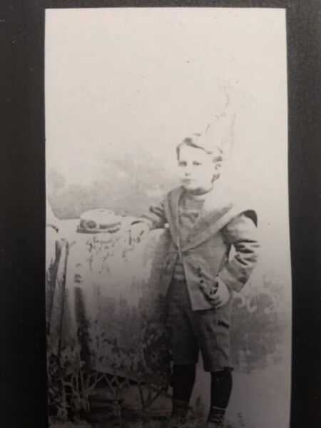 Black and white portrait of a young boy in vintage clothing, standing beside a table with a hat, showcasing late 19th-century fashion and photography style.