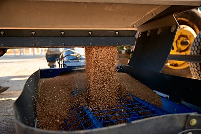 Two large trucks parked in front of several white agricultural silos under a clear blue sky, with a conveyor system extending towards the silos for grain handling.